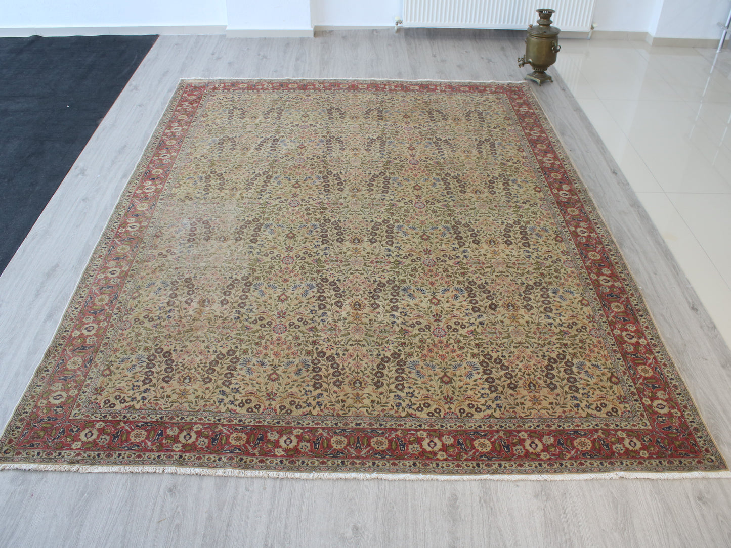 A rectangular traditional area rug with an intricate floral and geometric pattern, featuring a beige center field and a reddish-brown border, displayed on a light gray wood floor.