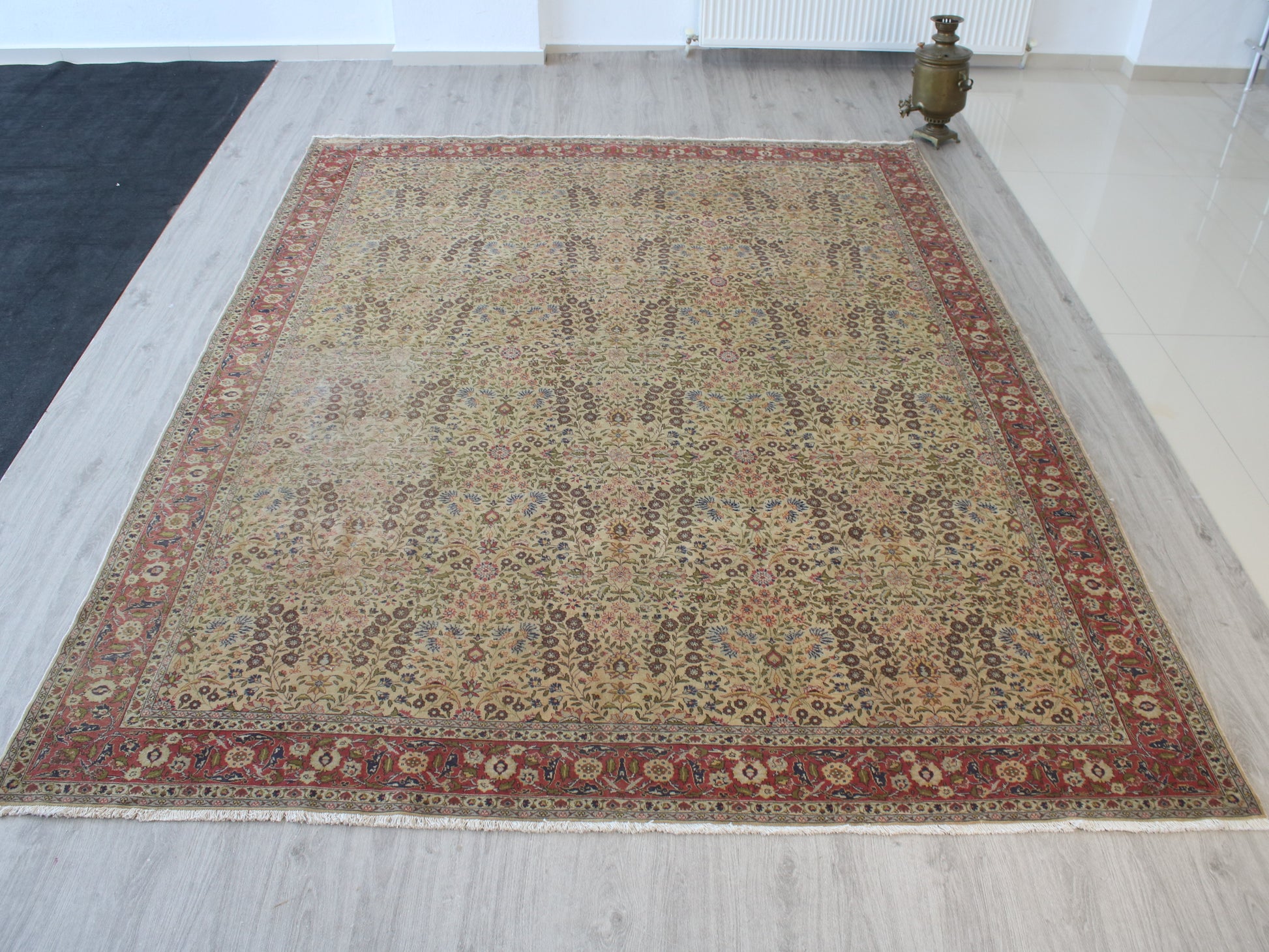 A rectangular traditional area rug with an intricate floral and geometric pattern, featuring a beige center field and a reddish-brown border, displayed on a light gray wood floor.