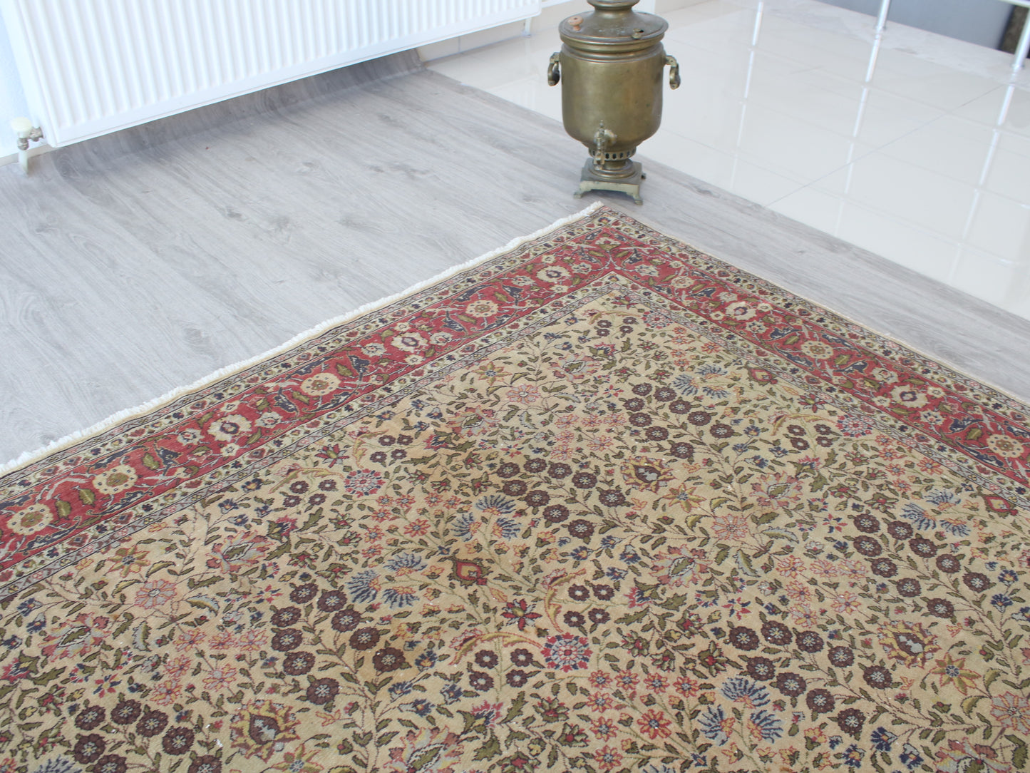 A traditional Persian-style area rug with an intricate floral pattern in beige, red, blue, and brown, shown on a light gray wood floor next to a brass samovar.