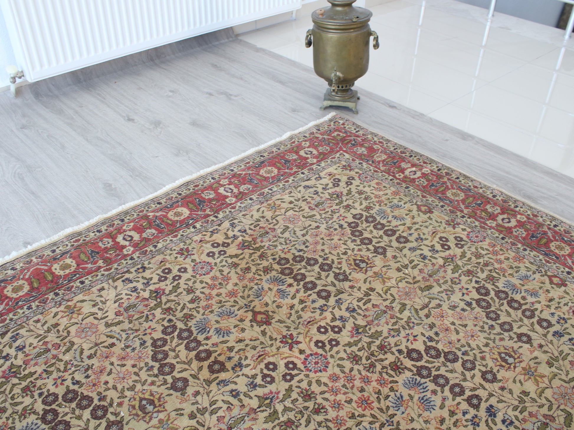 A traditional Persian-style area rug with an intricate floral pattern in beige, red, blue, and brown, shown on a light gray wood floor next to a brass samovar.