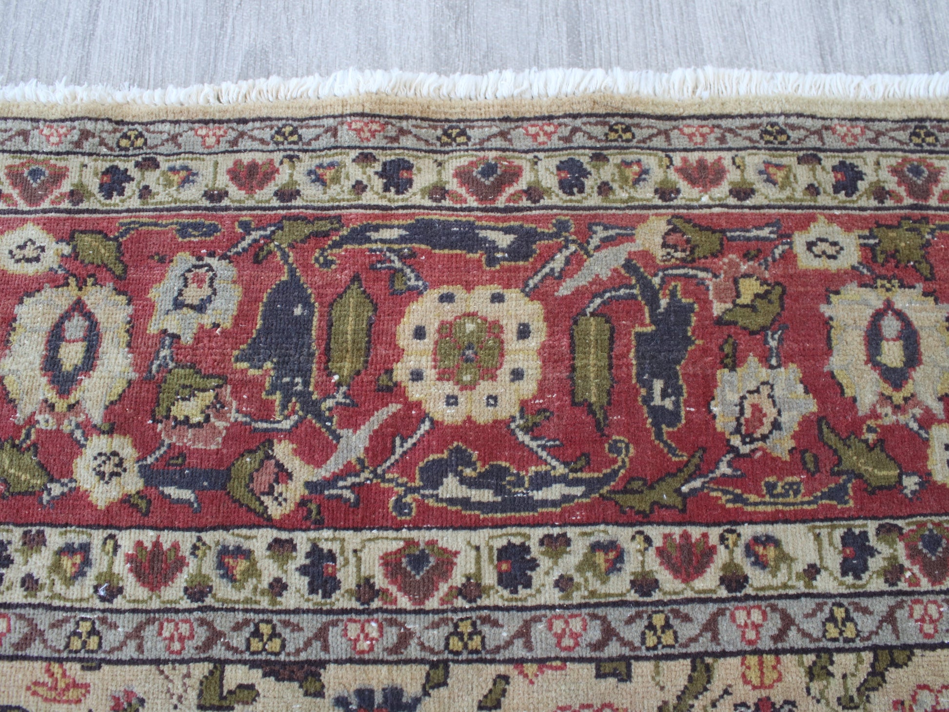 A close-up of a patterned area rug with a red field, cream borders, and multicolored floral motifs, showing white fringe at the top.
