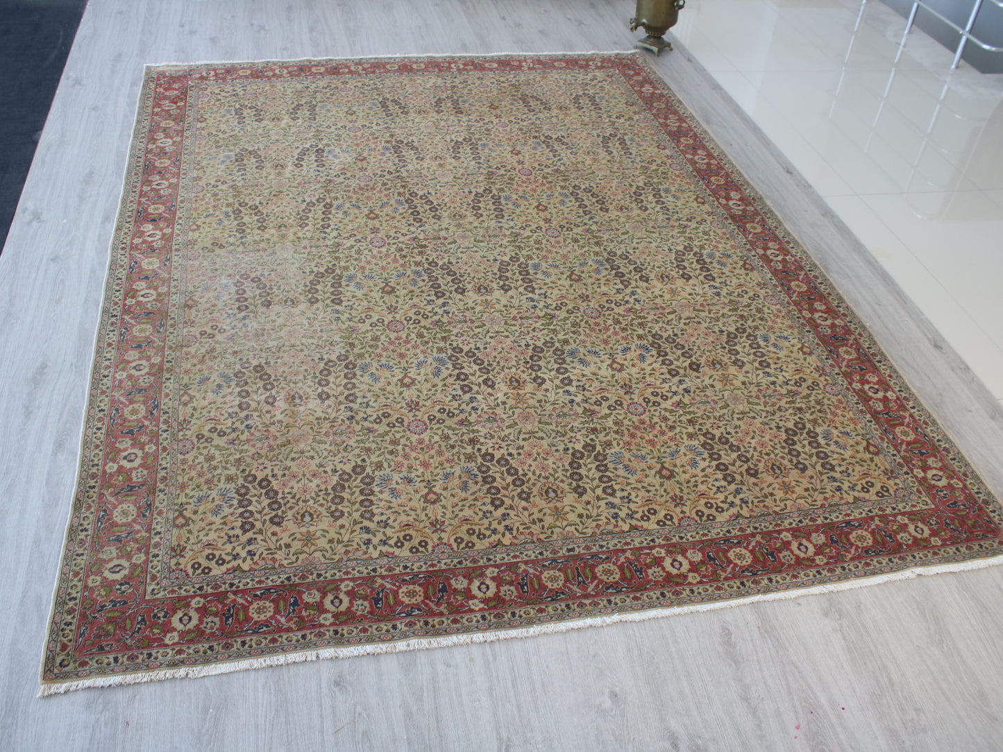 A rectangular traditional area rug with an intricate floral pattern, featuring a beige center field, a red border, and cream fringe, displayed on a light gray wood floor.