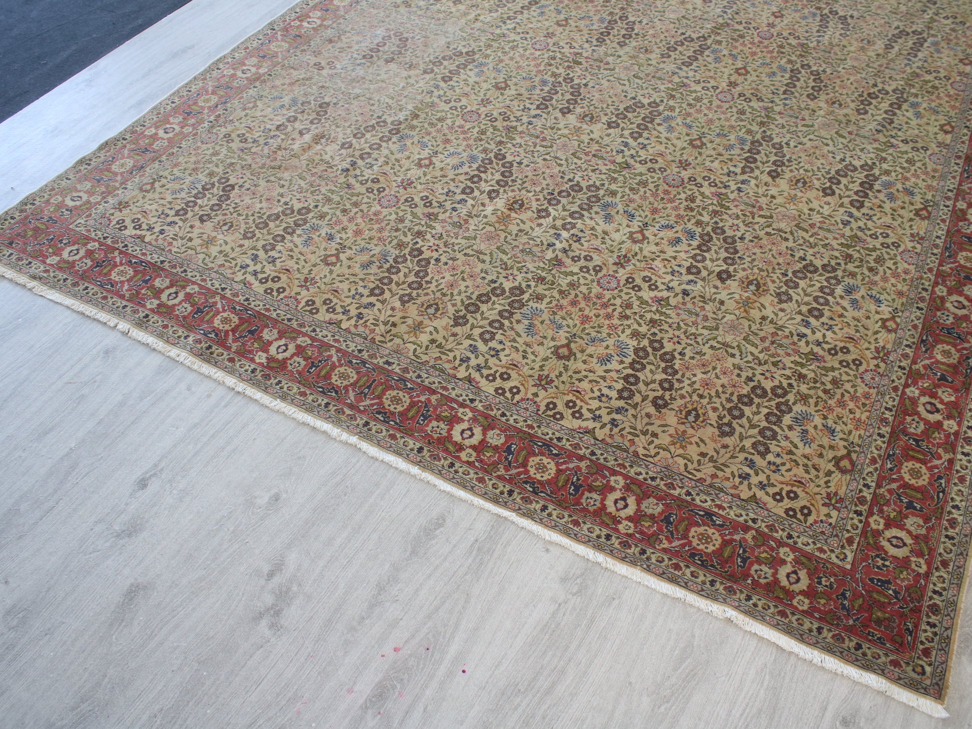 A traditional oriental area rug with an intricate floral pattern, featuring a beige center field and a red border, shown laid out on a light gray wood floor.