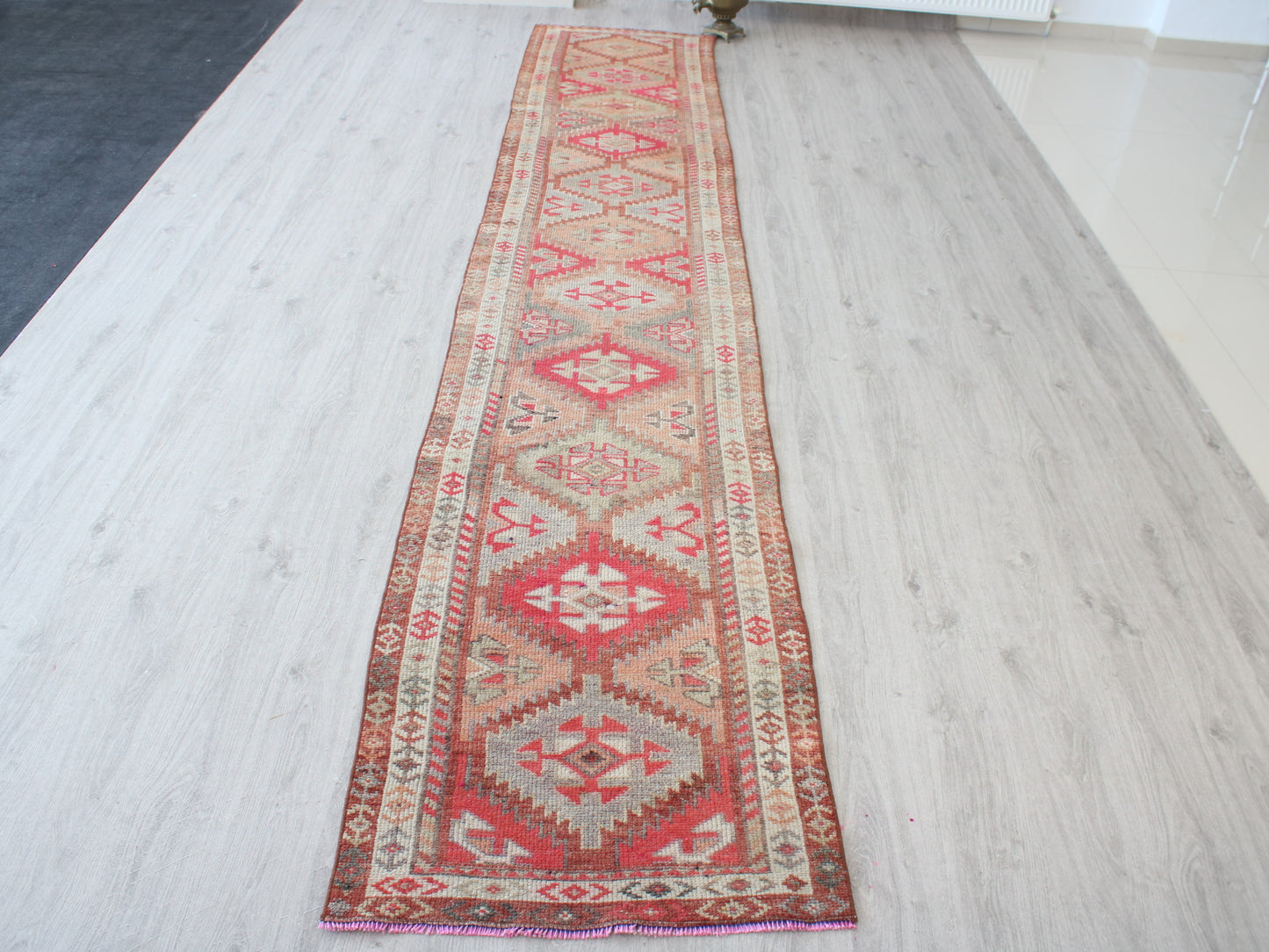 A long rectangular area rug with a traditional geometric pattern in red, beige, and brown, laid out on a light-colored wood floor.