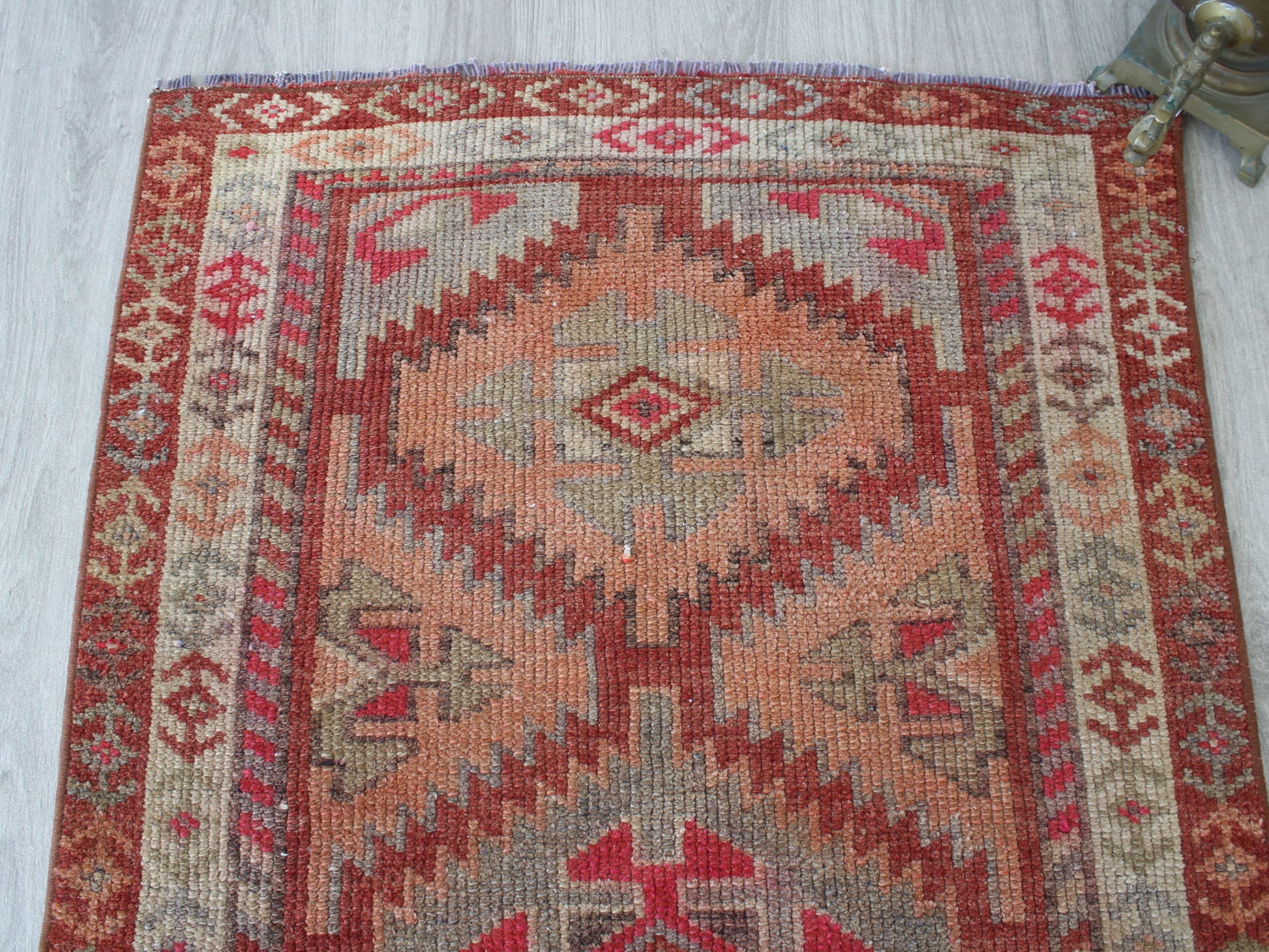 A square kilim-style area rug with a geometric diamond pattern in terracotta red, rust, beige, and gray, laid flat on a light wood floor.