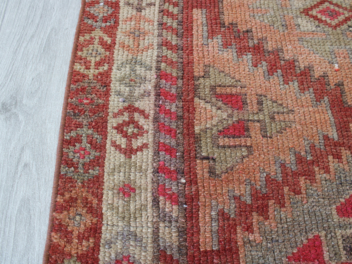A close-up of a patterned area rug with red, beige, and brown geometric designs, laid out on a light gray wood floor.