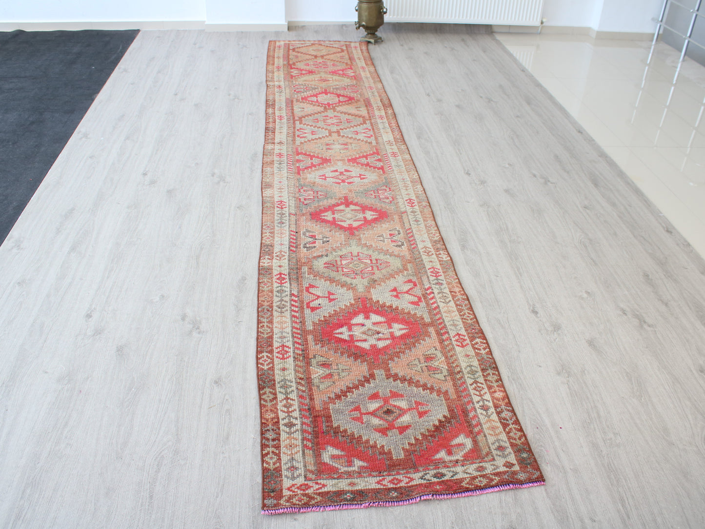 A long rectangular area rug with a traditional geometric pattern in red, beige, and brown, laid out on a light wood floor.