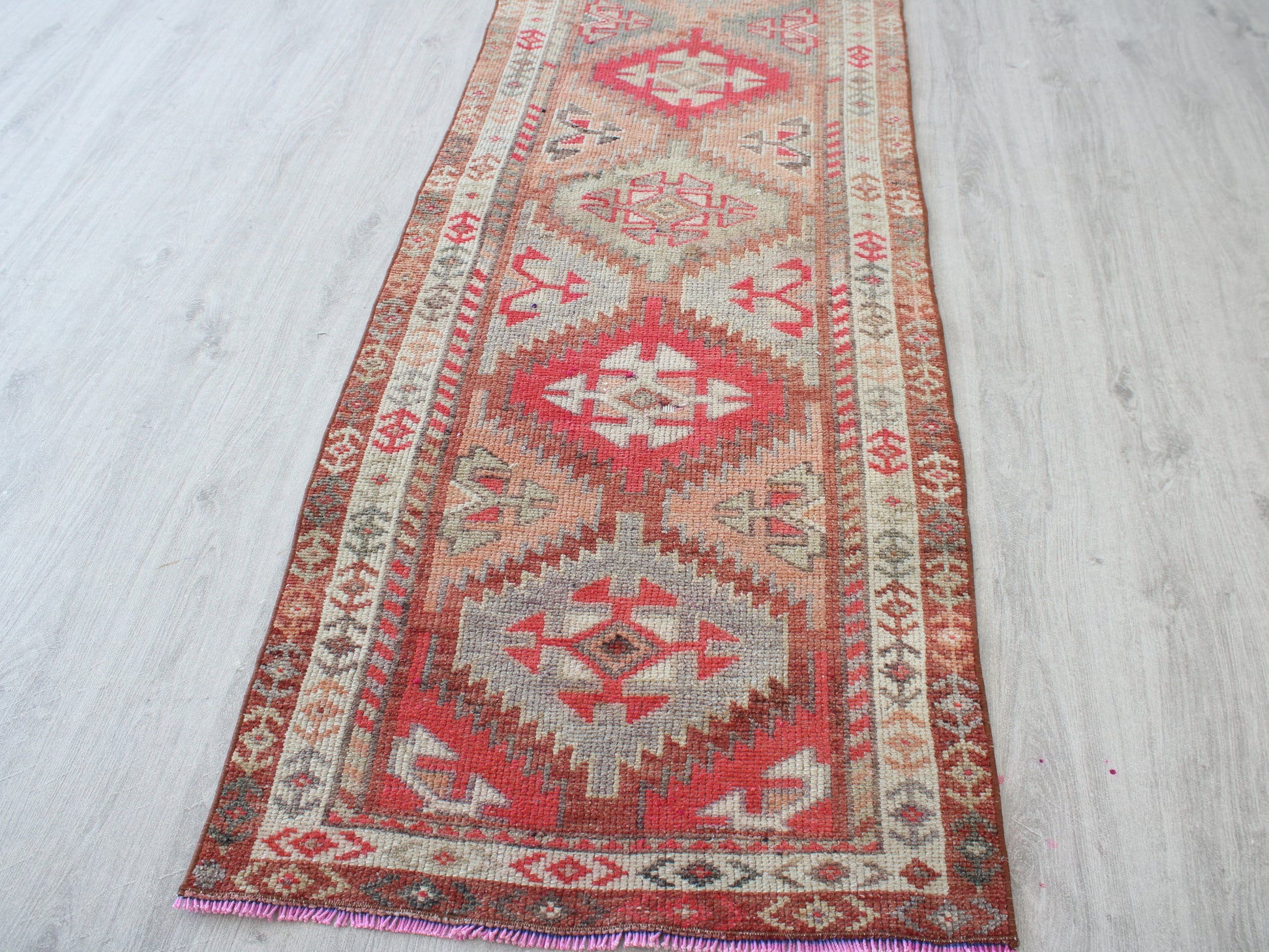 A long rectangular area rug with a traditional geometric kilim pattern, laid out on a light gray wood floor. The rug features red, pink, beige, and brown motifs.