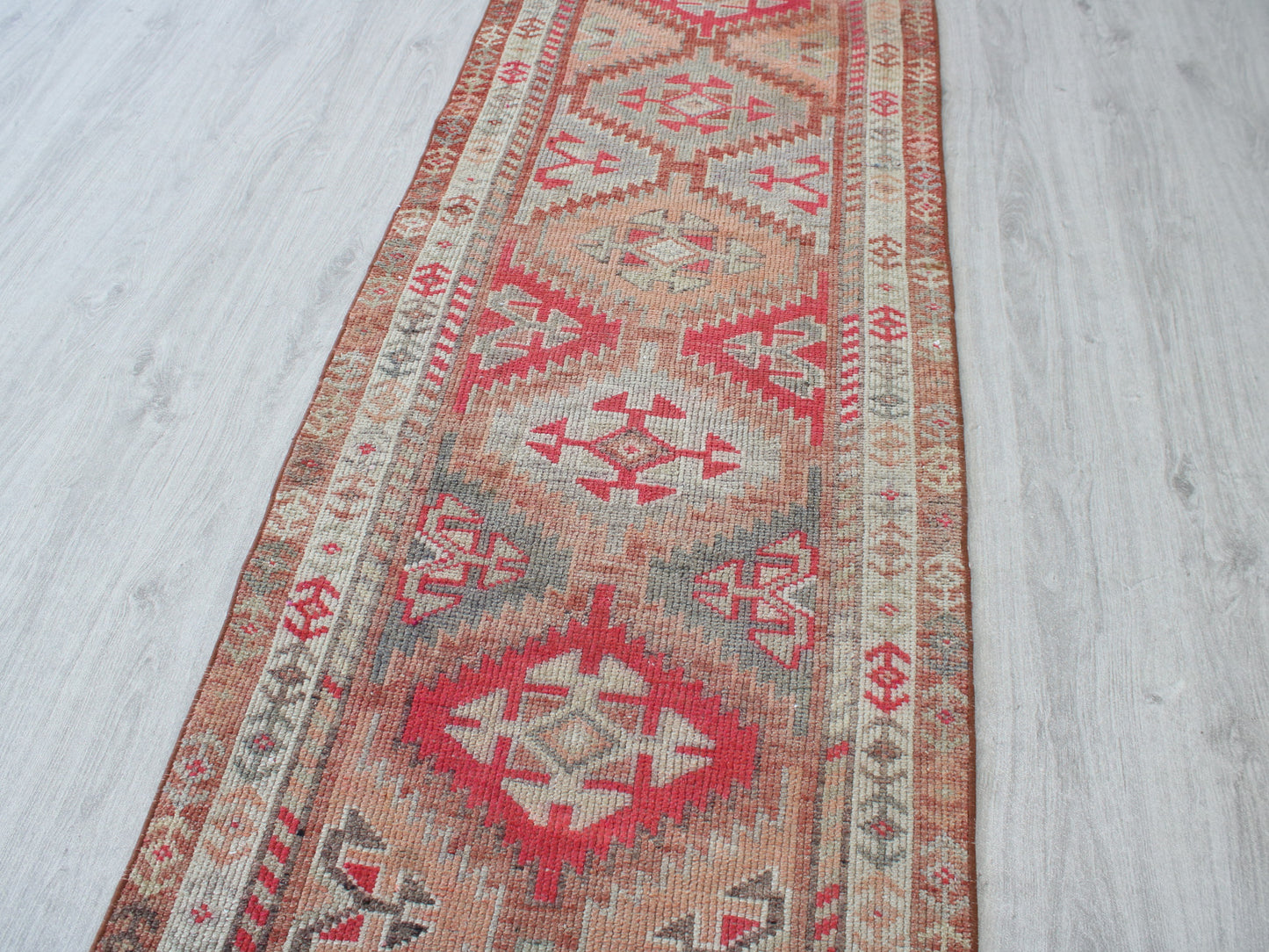 A long rectangular area rug with a distressed geometric tribal pattern in red, beige, and gray, laid out on a light gray wood floor.