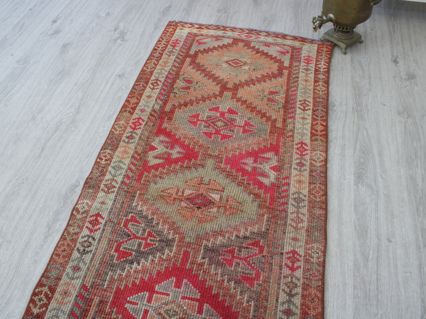 A long rectangular kilim runner rug with geometric patterns in red, beige, and brown, laid out on a light gray wood floor next to a brass object.