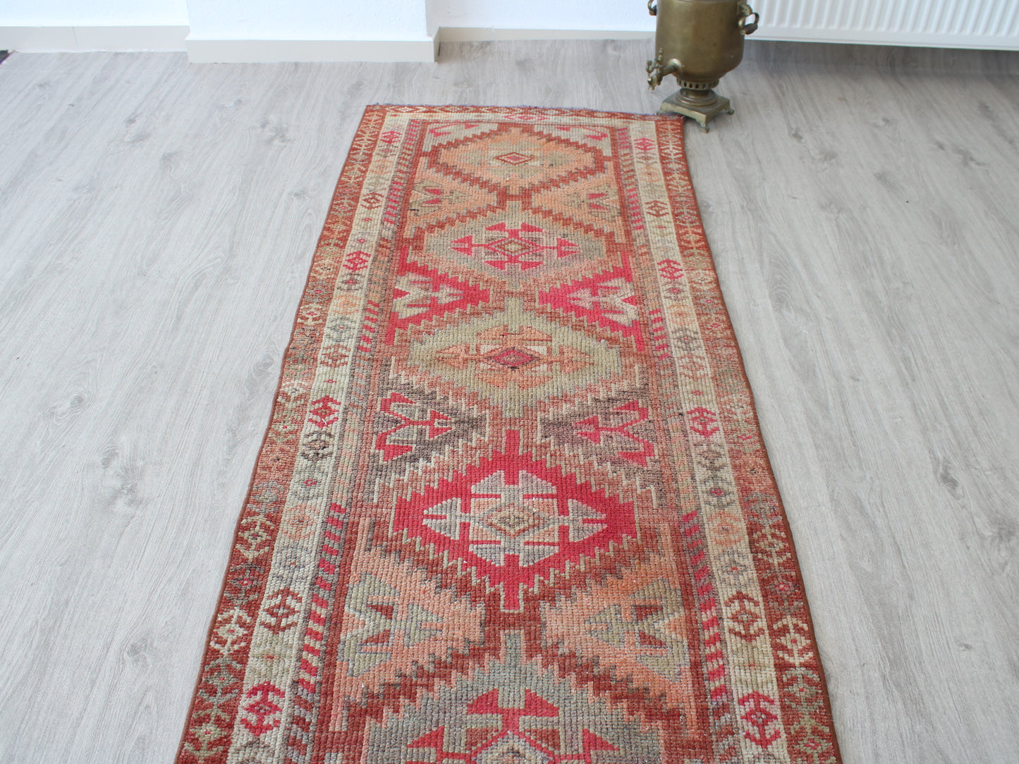 A long rectangular kilim runner rug with a distressed geometric pattern in red, pink, beige, and brown, laid out on a light gray wood floor.