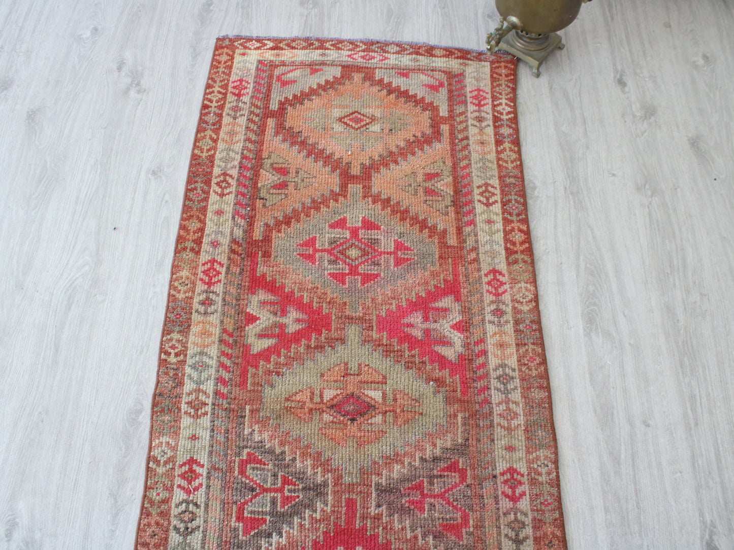 A rectangular vintage kilim runner rug with a geometric tribal pattern in red, beige, and brown, laid out on a light gray wood floor next to a brass lamp base.
