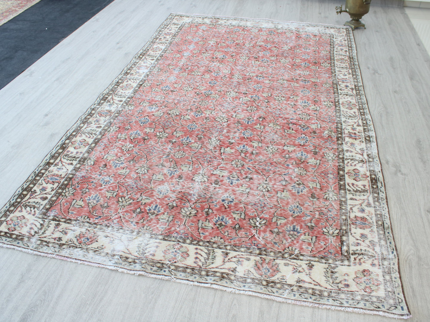 A rectangular vintage-style Persian rug with a faded red center and an ornate cream-colored border, laid out on a light gray wood floor.
