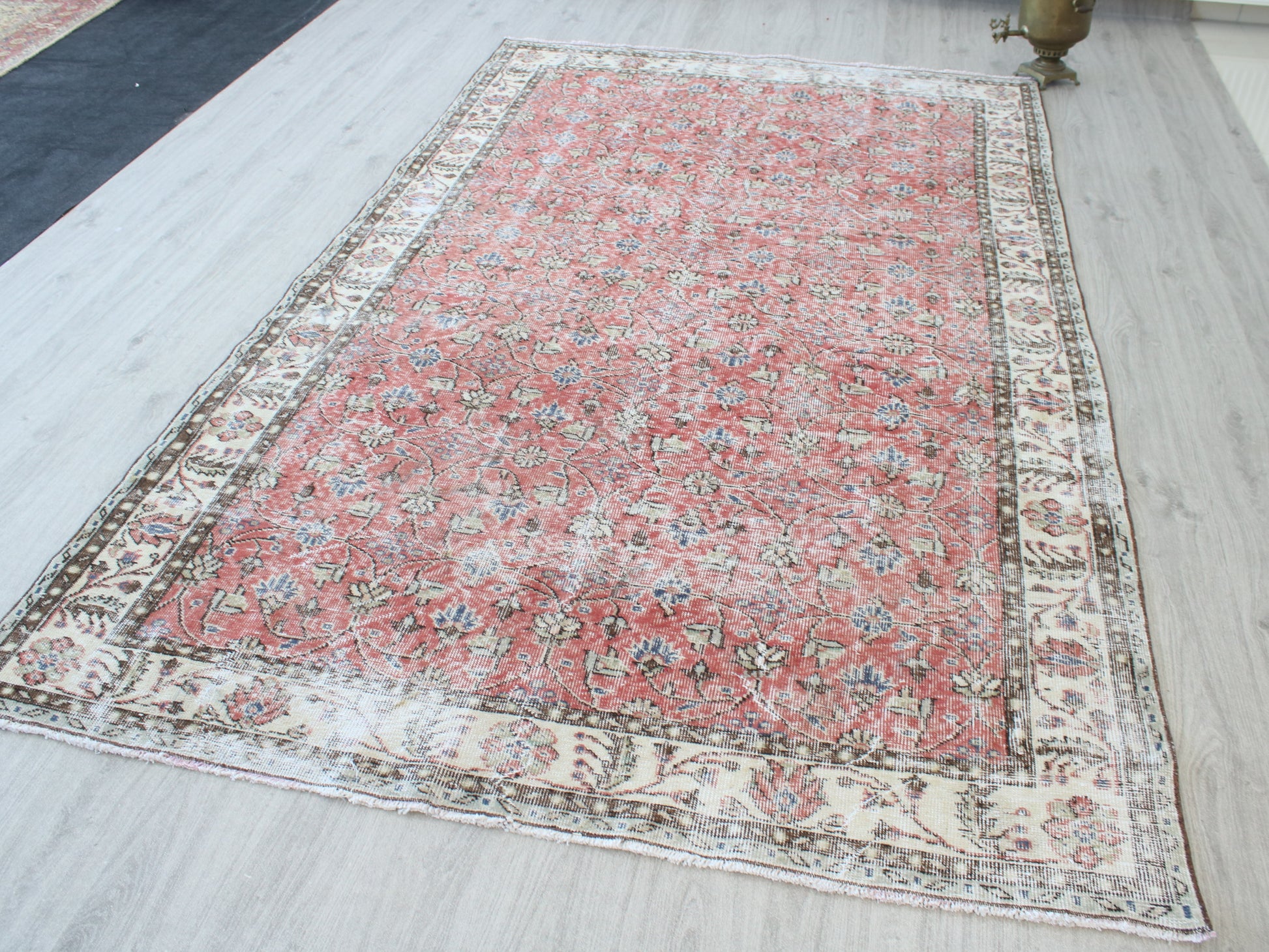 A rectangular vintage-style Persian rug with a faded red center and an ornate cream-colored border, laid out on a light gray wood floor.