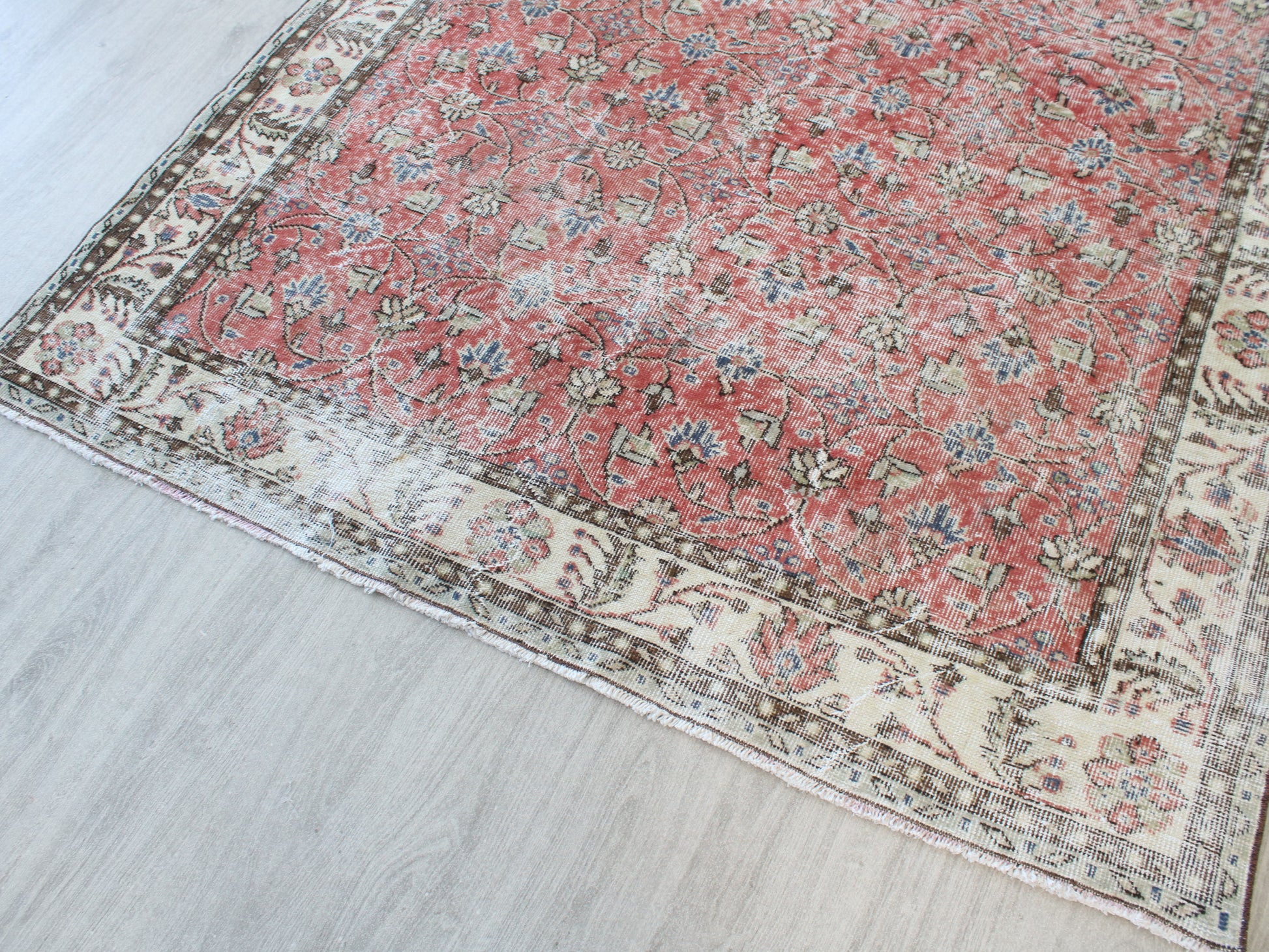 A traditional Persian-style area rug with a dusty red central field and an intricate cream-colored border, laid out on a light gray wood floor.