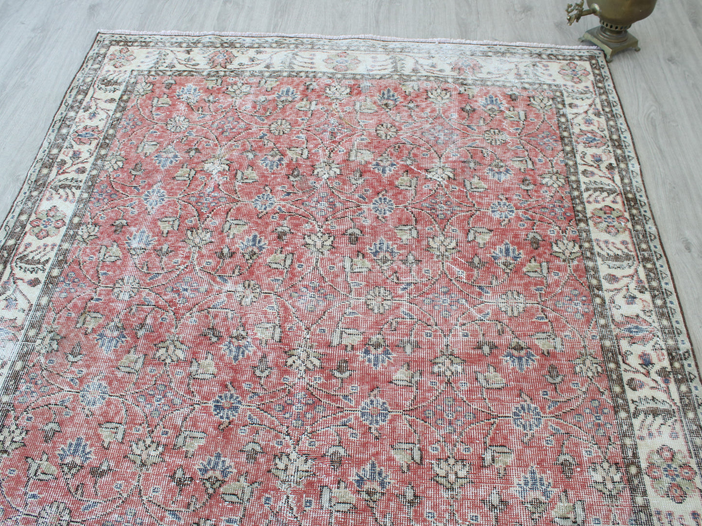 A rectangular vintage-style area rug with a distressed floral pattern, featuring a dusty red center field and a cream-colored border, laid out on a light gray wood floor.