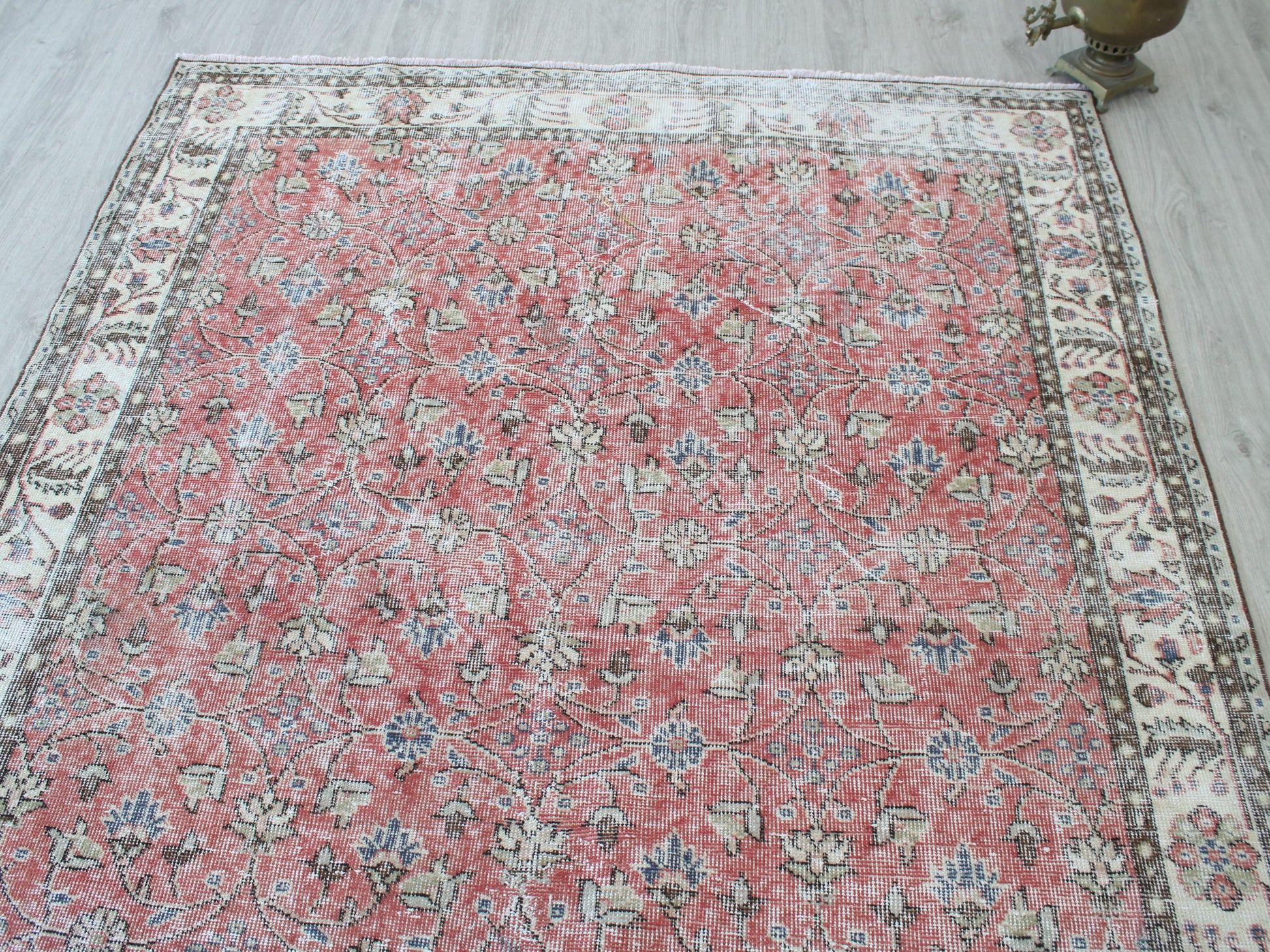 A rectangular vintage-style area rug with a distressed floral pattern, featuring a dusty red center field and a cream-colored border, laid out on a light gray wood floor.
