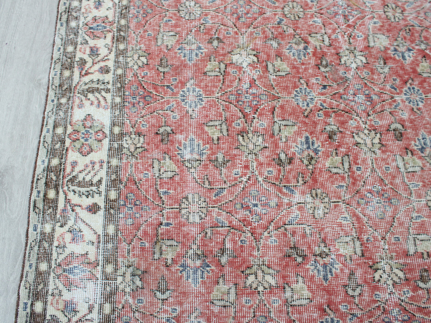 A close-up of a traditional patterned rug with a dusty red field, beige and cream floral motifs, and a detailed border on the left side.