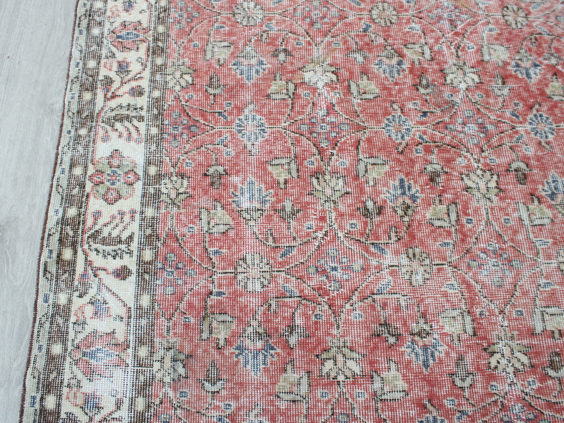 A close-up of a traditional patterned rug with a dusty red field, beige and cream floral motifs, and a detailed border on the left side.