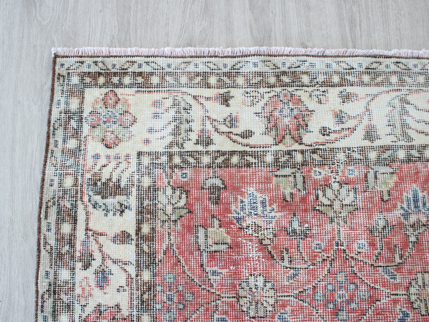 A close-up corner view of a traditional patterned rug laid on a light wood floor. The rug has a dusty pink central field with cream, brown, and muted blue floral motifs, and a matching cream border.