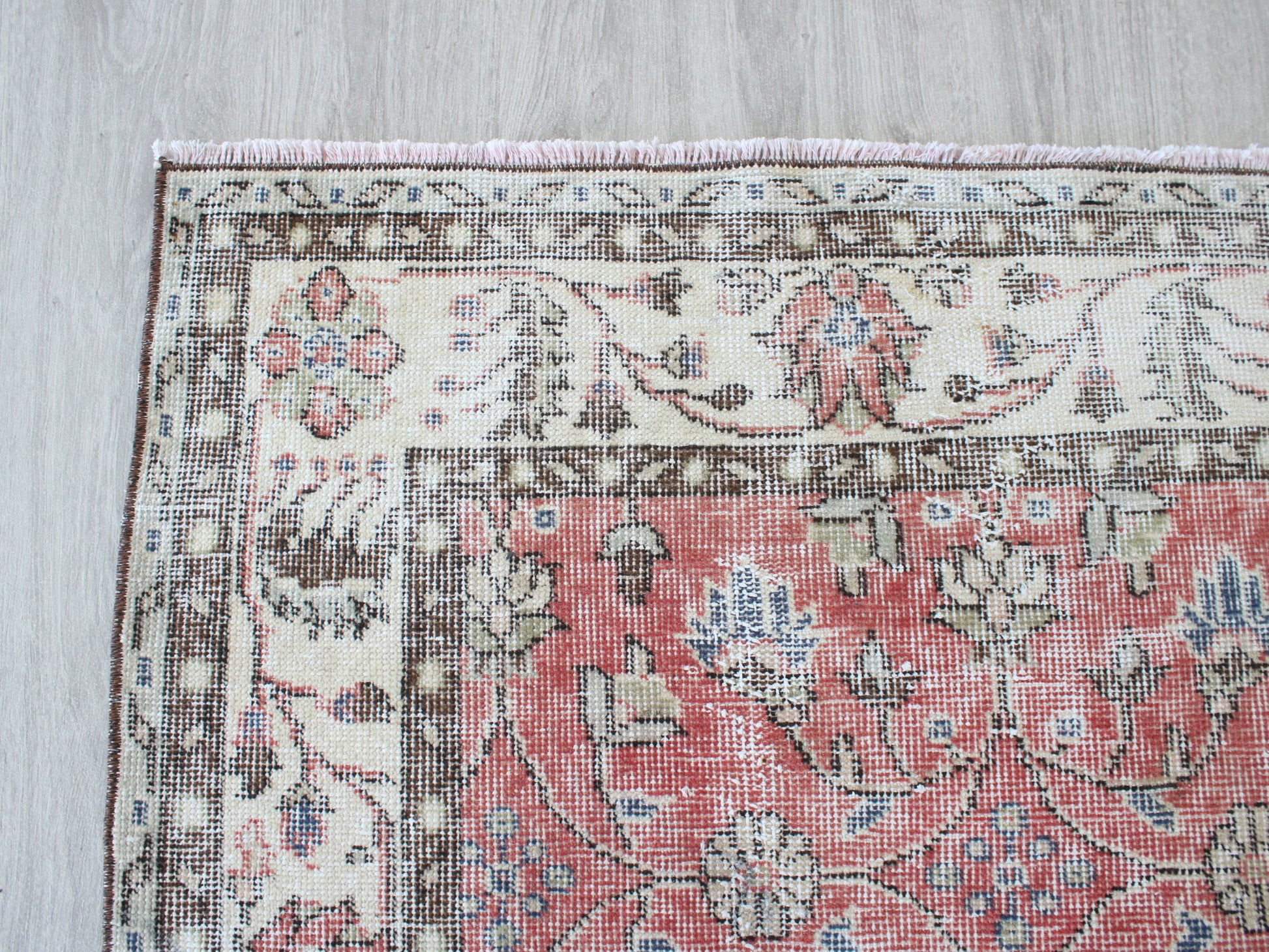 A close-up corner view of a traditional patterned rug laid on a light wood floor. The rug has a dusty pink central field with cream, brown, and muted blue floral motifs, and a matching cream border.