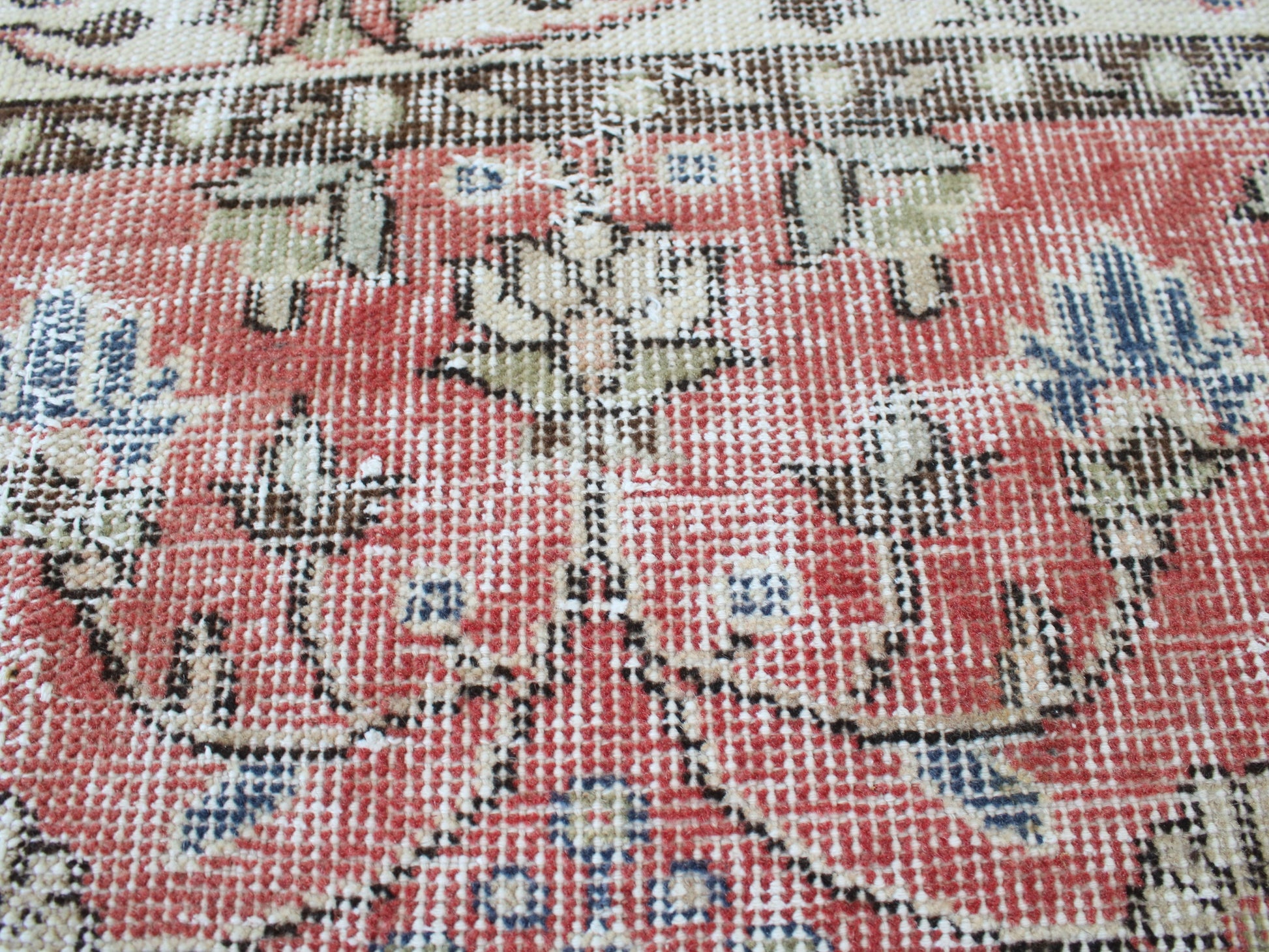 A close-up of a woven rug or textile with an ornate floral pattern in muted coral red, beige, blue, and brown tones.