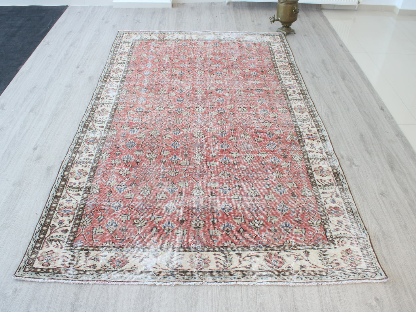 A rectangular vintage-style Persian rug with a distressed red center and ornate cream borders, laid out on a light wood floor.