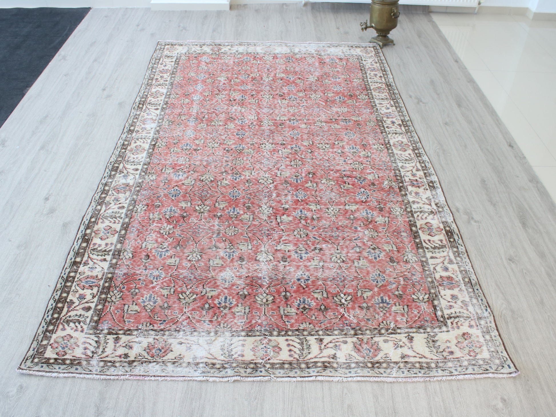 A rectangular vintage-style Persian rug with a distressed red center and ornate cream borders, laid out on a light wood floor.