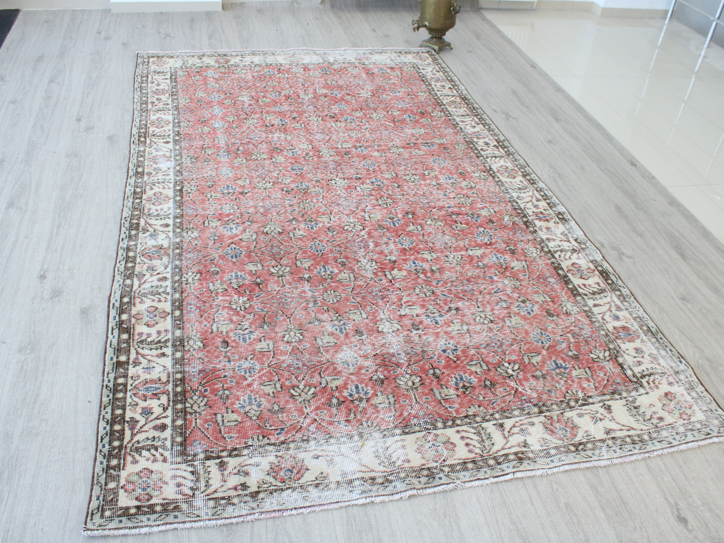 A rectangular Persian-style area rug with a distressed red center field and ornate cream, beige, and brown borders, displayed on a light gray wood-look floor.