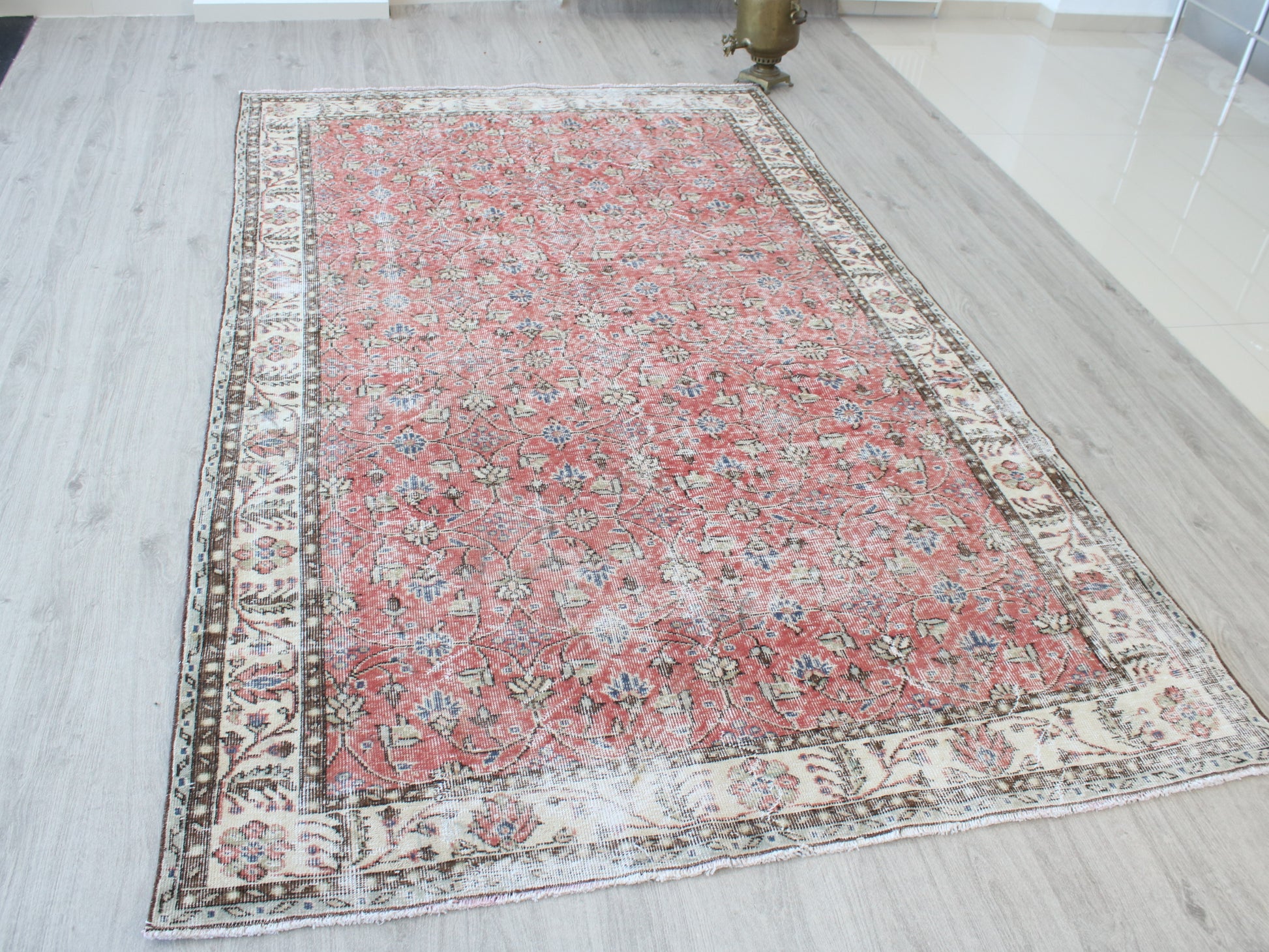A rectangular Persian-style area rug with a distressed red center field and ornate cream, beige, and brown borders, displayed on a light gray wood-look floor.